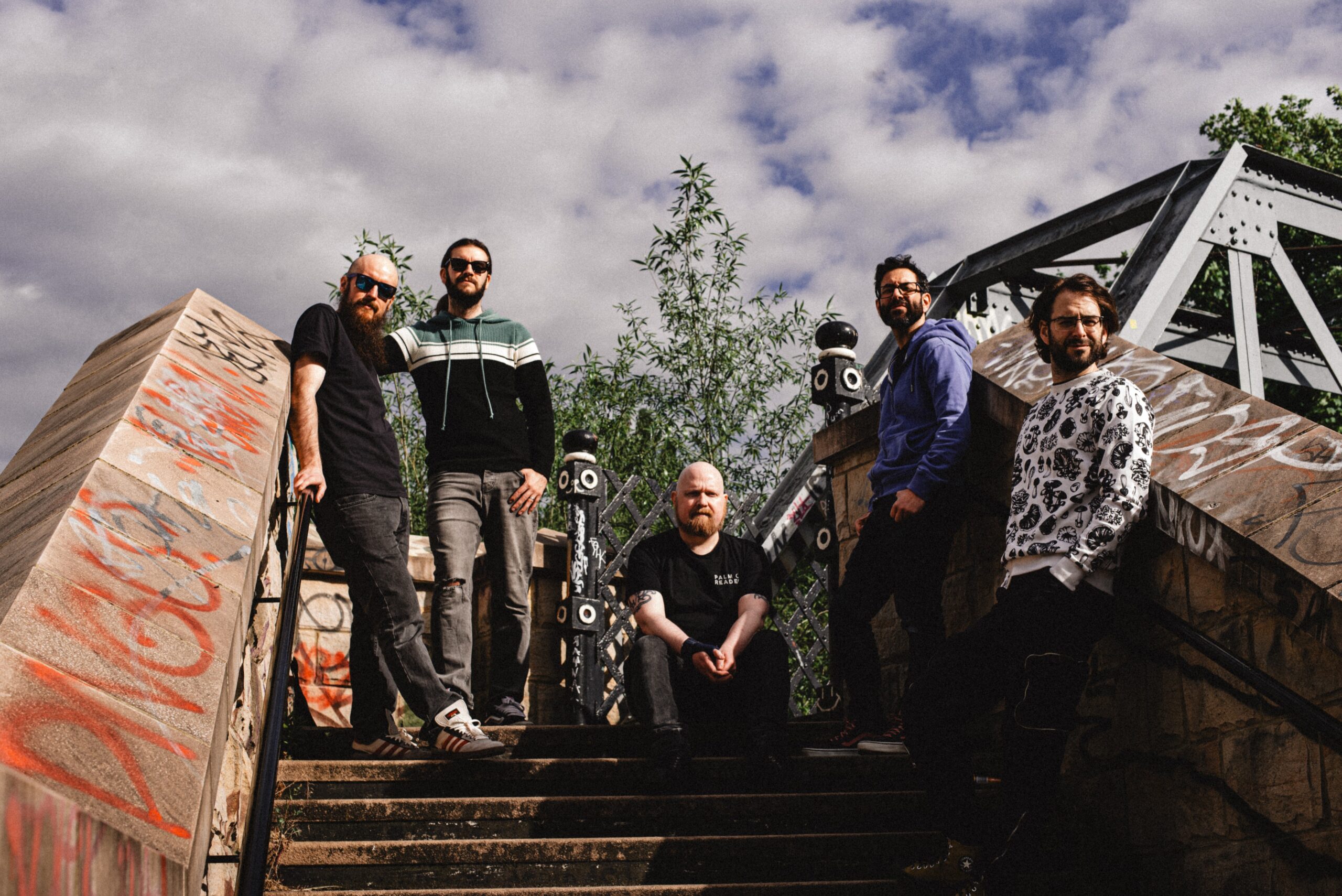The band Din of Celestial Birds. The five members of the band are stood on some stairs with a could sky in the background